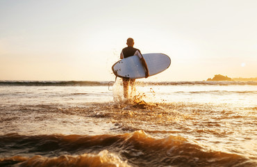 Surfer runs with surfboard towards ocean waves ta sunset time