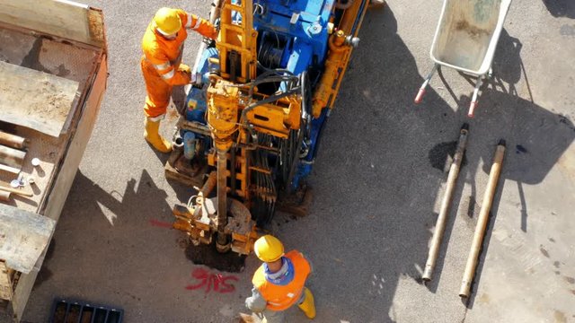 Two hydraulic workers operating powerful drilling rig in the centre of the square. Men using functional buttons and fixing long metal pipe for taking water samples for industrial explorations