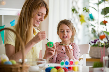 Obraz na płótnie Canvas A mother and daughter celebrating Easter, painting eggs with brush. Happy family smiling and laughing. Cute little girl in bunny ears preparing the holiday.