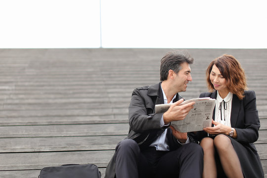 Engineers Male And Female Find Article About Company In Newspaper, Colleagues Sitting On Stairs Reading. Americans Wearing Business Clothes Smiling Communicating. Concept Of Fashionable Cl