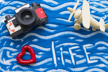 Life inscription on a blue colored sand with waves, roll film camera and wooden plane, top view, flat lay