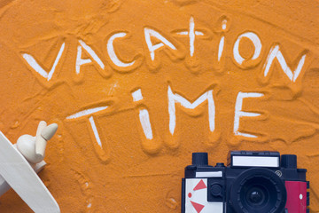 Vacation time inscription on an orange colored sand with roll film camera and wooden toy plane, top view, flat lay