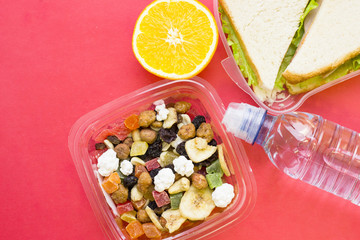Fresh sandwich with lettuce in a plastic container, a bottle of still water, orange and a plastic container of candied fruit, top view, rose background with copy space