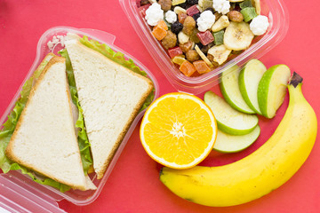Fresh sandwich with lettuce , orange, banana, slices of green apple and a plastic container of candied fruit, top view, rose background with copy space