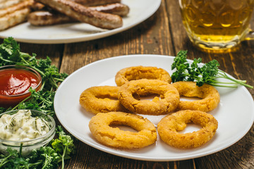 Delicious cheese rings and sausages on a white round plate with ketchup, sauce, parsley and beer