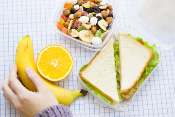 Fresh sandwich with lettuce in a plastic container, orange, a container of candied fruit and nuts and a man's hand holding a banana, top view, white squared paper background