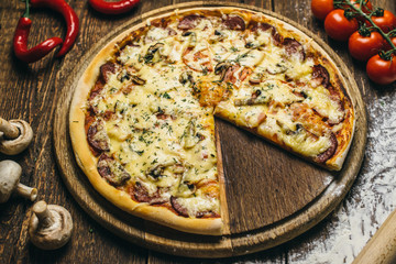 Delicious pizza with mushrooms, salami, cheese, tomatoes and pepper on a round wooden board on an old table. Top view