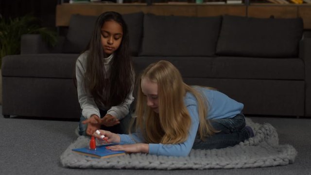 Cute little blonde girl doing manicure with red nail polish while her adorable mixed race girlfriend looking at beauty nails feeling happy. Happy preteen kids applying makeup in domestic interior.