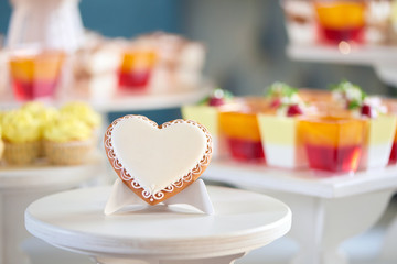 Close-up of little candy sweatheart,covered with white glaze and decorated with pattern, which stands on the wooden stand, near a yellow cupcakes and candybar on the background. Good gift for guests.