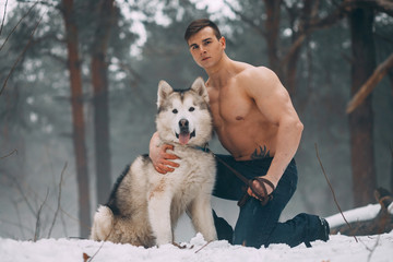 Young bodybuilder with bare torso embraces dog Malamute at walk in winter forest.