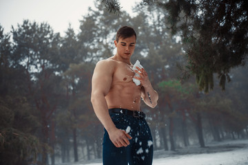 Young bodybuilder with bare torso wipes his body by snow in forest in winter.