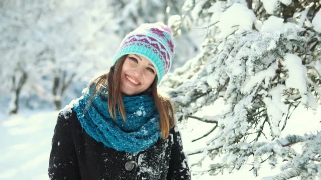 Young happy girl wears warm hat and gloves having fun before snowy christmas tree in the park, outdoor sunny day slowmotion