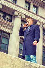 Fototapeta na wymiar American teenage college student traveling in New York, wearing blue long woolen overcoat, scarf, jeans, standing on street by vintage office building in winter, talking on cell phone. Filtered effect