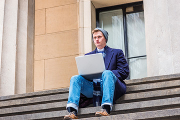 American college student traveling, studying in New York, wearing blue long woolen overcoat, scarf, jeans, leather shoes, knit hat, sits on stairs outside on street,  working on laptop computer..