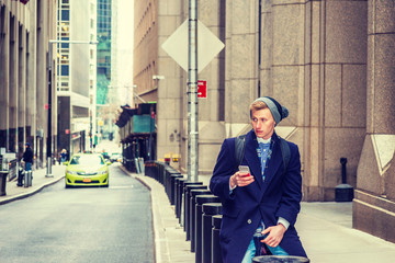 American man traveling in New York, wearing blue long overcoat, scarf, cuffed knit beanie hat, carrying back bag, sitting on vintage street, texting on cell phone. A small car on background..