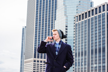 Young American man traveling in New York, wearing blue long woolen overcoat, scarf, cuffed knit beanie hat with pom pom, standing in front of business district, talking on cell phone. .