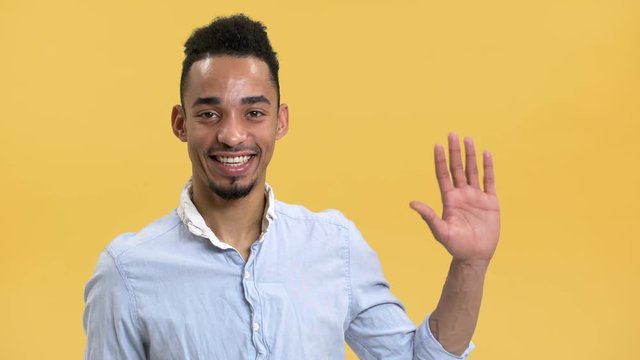 Portrait of happy arab guy with dark hairdo and mustache waving hand and welcoming, isolated over yellow background in studio. Concept of emotions
