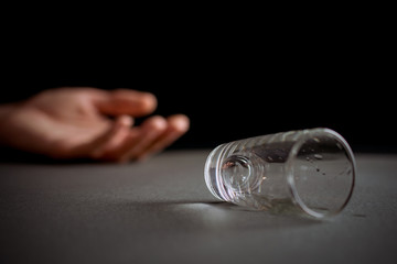lying empty glass of vodka or alcoholic drink, hand drunk in the background, concept of alcoholism and alcohol abuse, defocused, selective focus, close-up, gray table, dark background
