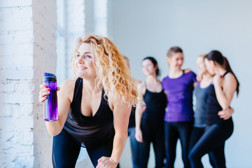 Attractive happy blond curly woman drinking water from the bottle in gym. Teamwork, good mood and healthy lifestyle concept.