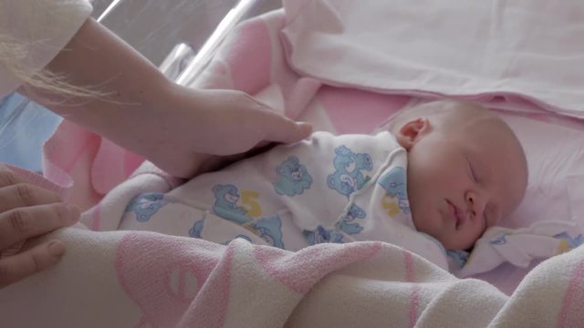 Mother stroking sleeping newborn baby lying in the bassinet in maternity hospital