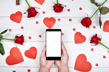 Close up of mobile smart phone with blank screen in woman hands, red paper hearts and rose flowers on wooden background, copy space. Flat lay, top view. Application mock up template