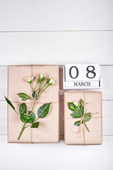Top view of gift boxes with branch of fresh roses and wooden cube calendar March 8 on white table, copy space. Holiday background, sale, shopping. Greeting card for Womens Day, flat lay
