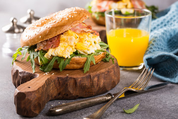 Sesame seeds Whole Grain bagels with scrambled eggs, rucola and fried bacon on small wooden chopping board on gray background. Healthy breakfast concept.