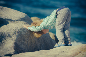 Boy standing on stone, playing on beach. Cute blond child leaned and peered into crevice of large stones on beach seaside coast. Flexible children's body, great gymnastics in nature. Yoga and sport