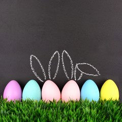 Row of Easter eggs in grass against a chalkboard background. Two with hand drawn Easter Bunny ears.