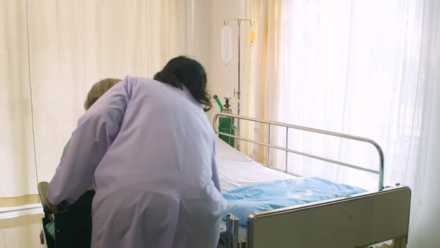 Professional female doctor helping her patient from wheelchair to the bed in hospital