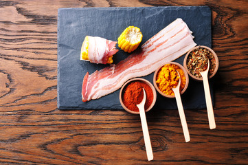 Bacon slices with corn cobs and wooden bowls with dry spices and spoons on the dark blue board on the wooden background