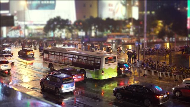 Cars and buses drive down the street with illuminated advertising and flashlights, people with umbrellas are walking along the road