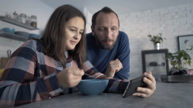 Father and daughter talking, eating and watching movie on smartphone
