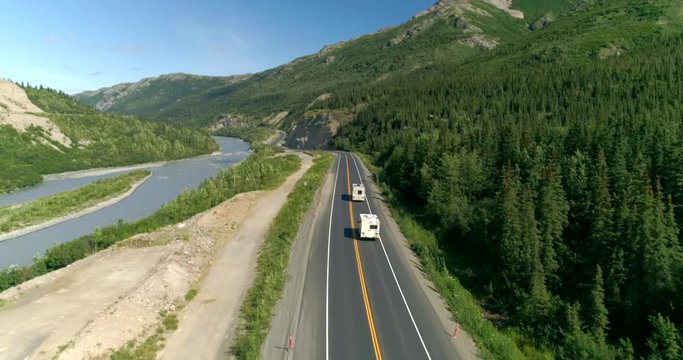 Aerial view of a motorhome and a pickup truck driving along an empty highway through a green river valley in Alaska. Spruce forest lines the road in a freeing adventurous scene of a summer road trip.
