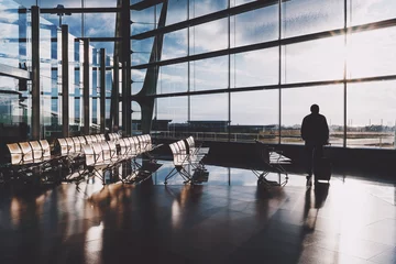Papier Peint photo Aéroport Silhouette of adult male tourist with his rolling bag standing alone in front of huge glass facade indoors of airport departure hall near empty rows of seats and waiting for his flight