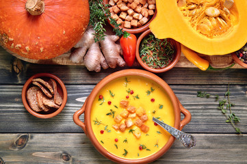 Pumpkin soup in ceramic bowl on rustic wood served with thyme and croutons. Next to it is a tray with healthy soup ingredients. Flat lay, view from above.