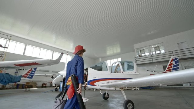 Female engineer walking to airplane and putting bag and tube on wing before repairing aircraft engine in hangar