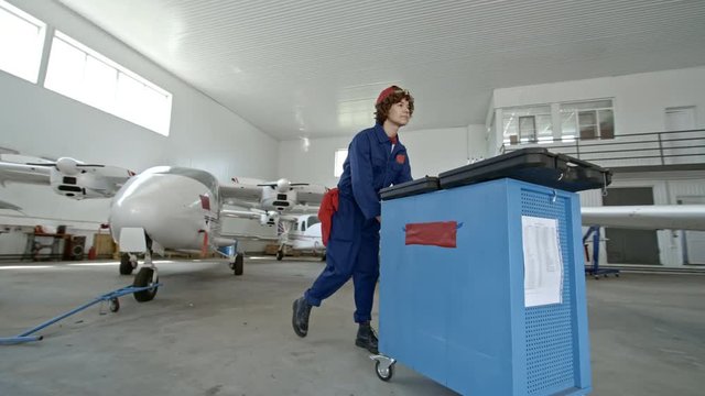 PAN of female engineer in uniform pushing cart with tools through hangar and inspecting airplane engine
