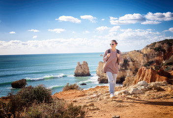 Naklejka na ściany i meble A traveler with a backpack walks along the rocky coast of the ocean, admiring the incredible scenery. Portugal, the Algarve, a popular destination for travel in Europe