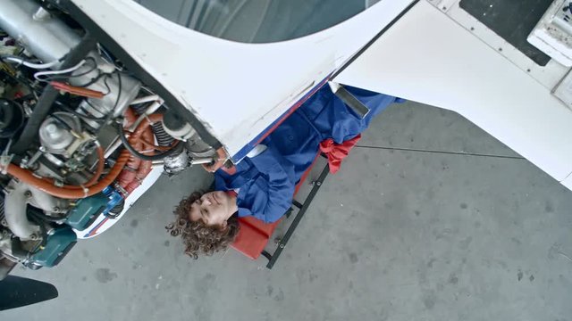 Top view of female aircraft mechanic lying on creeper underneath of airplane with exposed engine and working with screwdriver