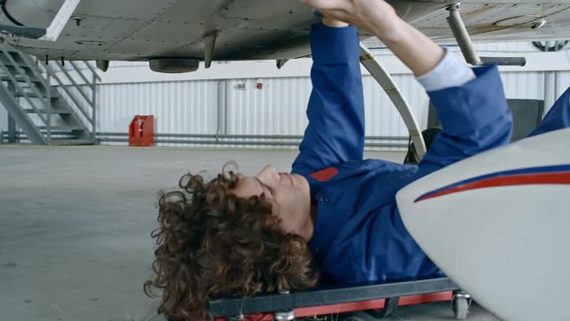 Female aircraft mechanic lying on creeper underneath of airplane and repairing vehicle with instrument