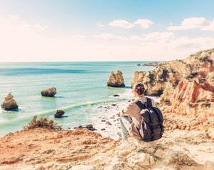 Fototapeta na wymiar Girl traveler with a backpack sits on the rocks on the ocean, admiring the incredible scenery. Portugal, the Algarve, a popular destination for travel in Europe