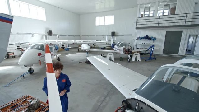 Tilt down of female maintenance engineer in uniform walking to cart with instruments, taking screwdriver and fixing airplane engine in hangar