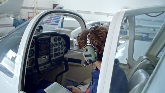 Tilt down of female pilot with headset sitting in cabin of jet airplane in hangar, using digital tablet and checking cockpit