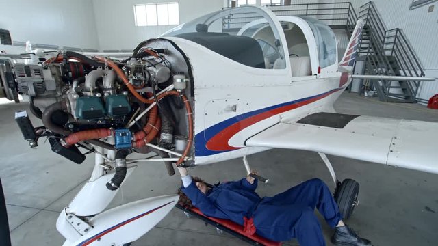 Tilt down of female aircraft mechanic lying on creeper underneath of jet airplane and cleaning bottom with towel