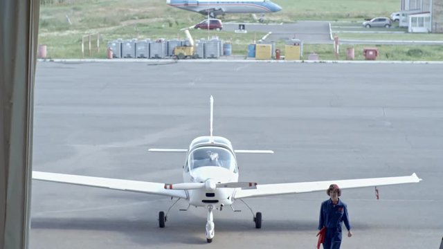 Tilt down shot of female aircraft mechanic walking into hangar from aerodrome with light jet airplane
