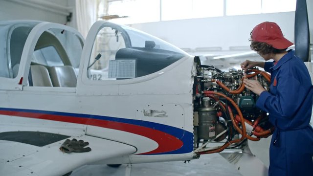 Tracking shot of young female aircraft mechanic repairing airplane engine in hangar and then turning at camera and happily smiling