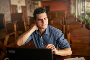 Businessman with backpain after long hours at work rubbing his neck because of ache. Overworked handsome freelancer man with scoliosis working with laptop. Health problems theme.