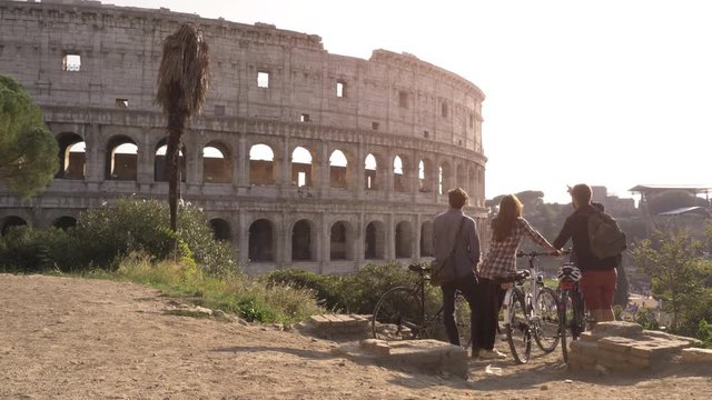 Three happy young friends tourists with bikes and backpacks at Colosseum in Rome standing on hill at sunset with trees slow motion steadycam wide shot