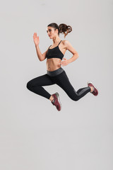 Full-length photo in profile of happy healthy woman in sportswear with abs jumping and flying in studio, isolated over gray background